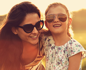 Obraz premium Happy fashion kid girl embracing her mother in trendy sunglasses and looking on nature background. Closeup toned color portrait of happiness.