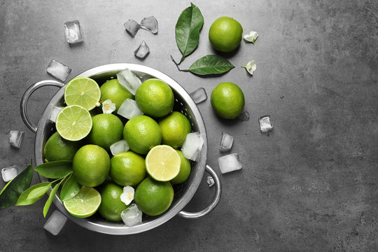 Colander with fresh ripe limes and ice cubes on gray background, top view