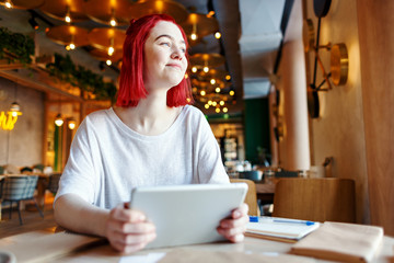 Inspired female student with dyed red hair sitting at table in cafe, holding tablet computer and looking through window