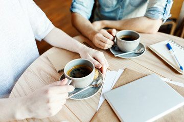 Hands of unrecognizable man and woman having coffee during business meeting in cafe, tablet computer, notepad and papers on table nearby