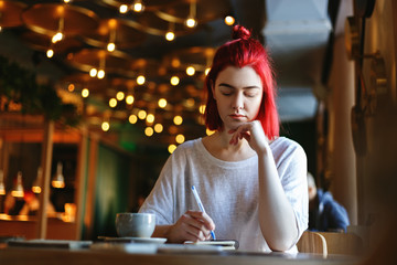 Portrait of young woman with red hair studying or working in cafe. Creative girl sitting at table...
