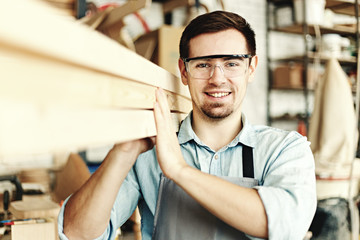 Young furniture maker carrying wood planks on his shoulder. Woodworker wearing protective...
