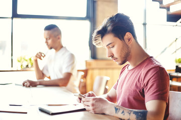 Profile view of handsome college student siting at desk and text messaging on cell phone, his fellow student in background