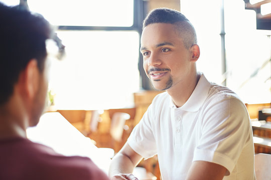 Mixed race young man talking to his unrecognizable friend while sitting at table in cafe