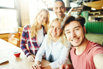 Four happy diverse university friends smiling at camera while taking selfie together in daylight