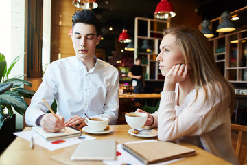 Young woman drinking coffee while her male colleague writing down notes in notebook in cafe