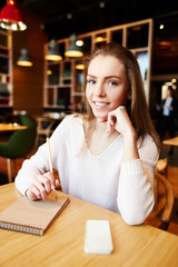 Content pretty woman sitting with phone and notepad at table in cafe smiling at camera