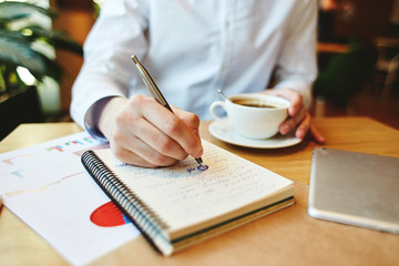 Close-up of unrecognizable man in white shirt having coffee while sitting at table and making calculations in notepad