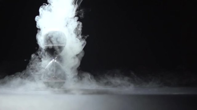 Smoke of liquid nitrogen pours on the glass hourglass in the frame.