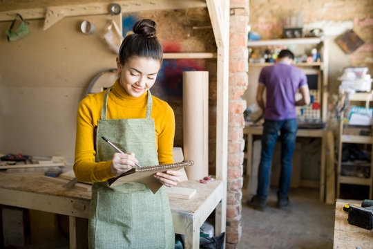 Waist up portrait of pretty young woman wearing apron holding notebook and drawing sketch in crafting shop, standing against brick wall, copy space