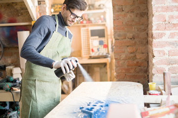 Portrait of modern carpenter spray painting wood piece while working in joinery workshop, copy space