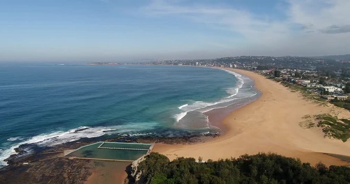 Headland of Narrabeen north beach with rock pool on sandstone rocky plato towards sandy wide beach and surfing activities.
