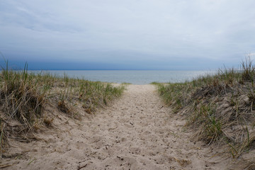 Fototapeta na wymiar Sand with beach grass in foreground, cloudy sky, lake in background