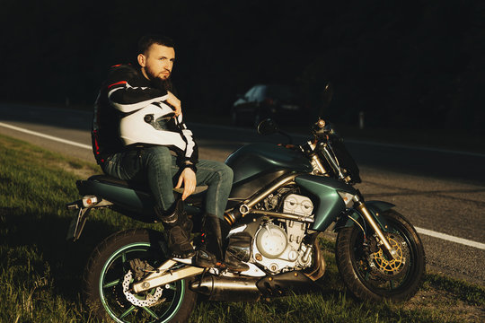 Full length portrait of a handsome serious biker sitting on his motorcycle with a hand on his helmet and looking away near the road against the forest.