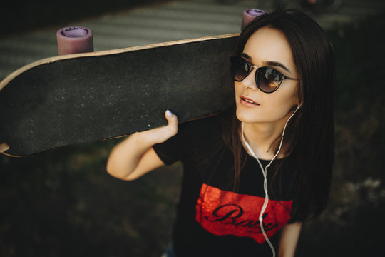 Close up portrait of a lovely caucasian girl holding a skateboard on her shoulders , wearing sunglasses and looking into camera smiling while listening music.