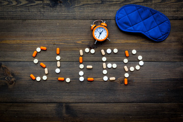 Medicine helps get asleep. Good sleep. Word sleep lined with sleeping pills near sleeping mask and alarm clock on dark wooden background top view