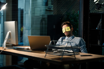 Office worker sleeping at table covering eyes with stickers having painted eyes. 