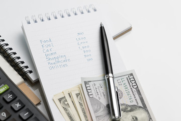 Expense, cost or budget calculation concept, hand writing list numbers of expenses annual cost on small paper white notepad with pen, calculator and US dollar bills on white table