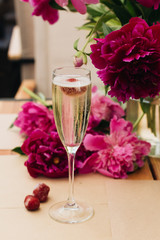 Glass of sparkling wine with cherry, summer drink, pink peony flowers on table