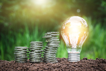 Light bulb with coins beside concept put on the soil in soft green nature background.