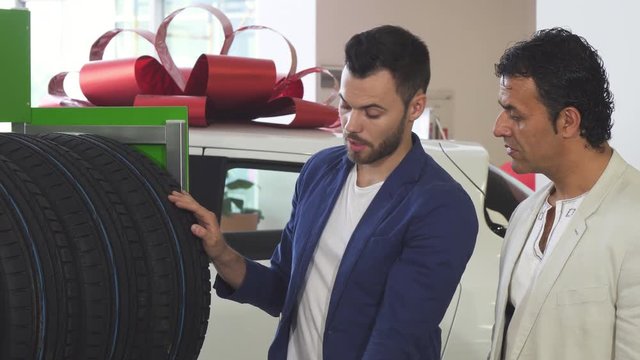 Handsome young professional car dealer talking to a mature man showing him tires for sale. Salesman working at the automobile dealership. Mature man buying tires for his auto.