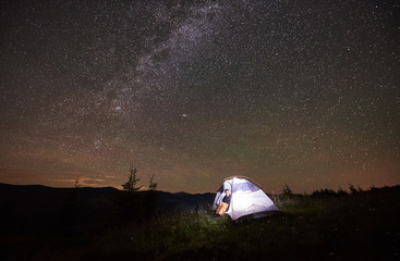 Fototapeta na wymiar Happy girl traveller having a rest at night camping in the mountains under incredible beautiful starry sky and Milky way. Smiling woman sitting inside illuminated tent and looking at sky full of stars