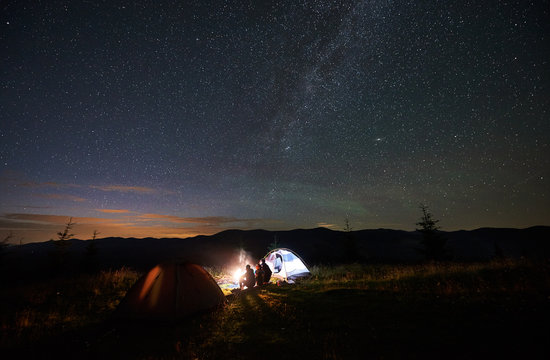 Back view of mother and two children tourists having a rest at night camping in mountains, sitting on log beside campfire and glowing tent, looking at beautiful starry sky and Milky way in evening