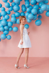 Fototapeta premium Happy smiling girl in cute white dress dancing on pink background. Modern studio decorated with blue shiny balls. Copy space for text.
