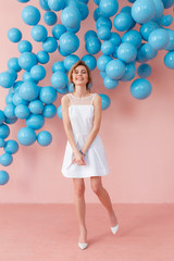 Fototapeta premium Happy smiling girl in cute white dress dancing on pink background. Modern studio decorated with blue shiny balls