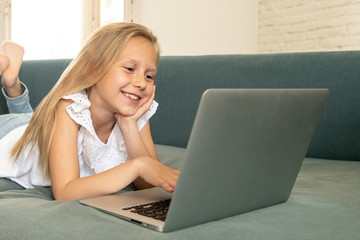 Beautiful cute charming little blonde girl playing and surfing the internet on laptop smiling and lying down on the couch at home. Digital technology, internet usage and life style concept