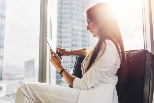 Successful Woman Wearing Suit Sitting On Armchair Using Tablet Computer At Her Loft Apartment In City Center