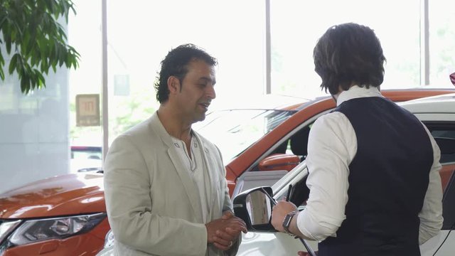 Mature handsome Hispanic man talking to the salesman at the car dealer. Male customer buying a new automobile at the dealership salon. Consumerism, lifestyle, transportation concept.