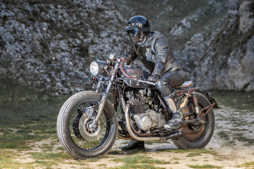 Biker with black leather suit and mask stay on his custom special rat motorbike.  Desolated rocks in the background. Post apocalyptic concept