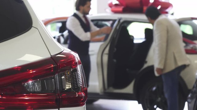 Selective focus on a car on the foreground. Professional automobile salesman working with his client on the background, opening car dor for a mature man. Male customer choosing new auto.