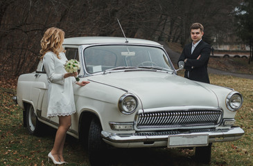 Just married wedding couple is standing near the retro vintage car in the park. Spring or autumn grey day in forest. Blonde bride in elegant white dress with bouquet and elegant groom in love.