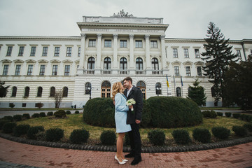 Obraz premium Just married wedding couple is standing near beautiful architecture building. Bride in blue coat and groom in grey. Couple is hugging outdoors on early spring or autumn day. Green bushes on background