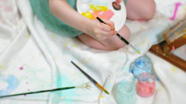 Beautiful red-haired girl sits on the floor in the studio in front of the easel and draws a brush and colored paints