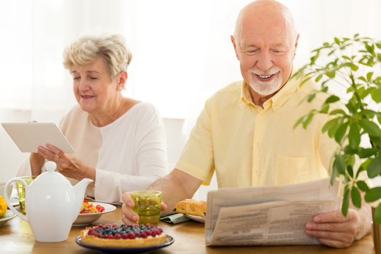Happy elderly woman using tablet while her husband reading newspaper