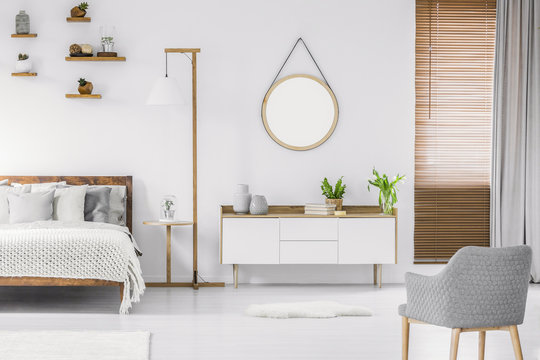 Scandinavian style white room interior with round mirror on the wall, wooden bed with pillows and blanket, cabinet and armchair. Real photo.