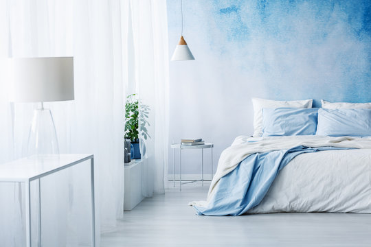 White lamp on a table in bright blue bedroom interior with bed against ombre wall