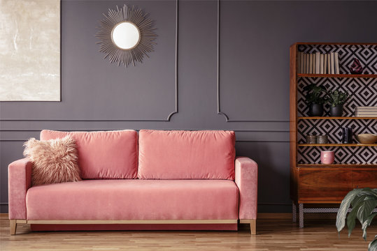 Simple, pink sofa with a fur pillow next to a wooden cupboard in living room interior with grey wall and mirror