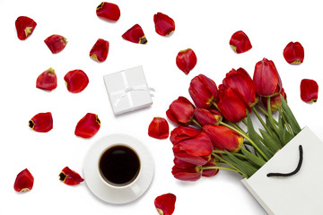 Fototapeta na wymiar Red tulips in a paper gift bag, gift, a cup of coffee and scattered petals of tulips on a white background. Top View.