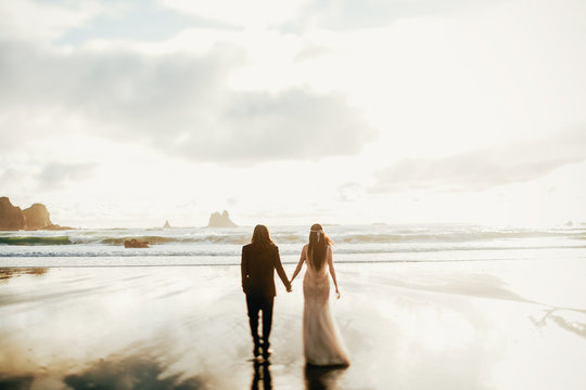 Adorable wedding couple holds each other hands walking along the beach against the rocks and ocean waves in the evening light