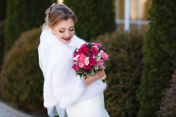 Bride in a white coat posing outdoors, holding a bright bouquet of flowers in her hands. The girl lowered her eyes and showed her make-up of natural tones.