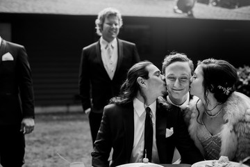 Bride and groom kiss a man while he hugs them at a dinner table