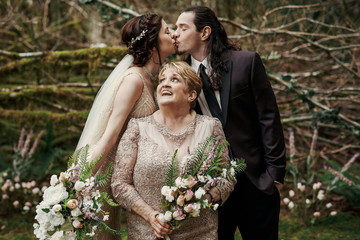 Bride and groom kiss behind smiling mom standing before them in mystic forest