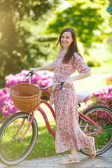 Portrait of smiling young woman in long pink floral dress stop to riding on vintage bike with...