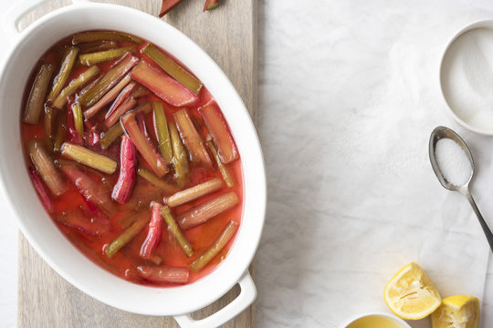 Cooked rhubarb in a white oval serving dish with a bowl of sugar and squeezed lemons.