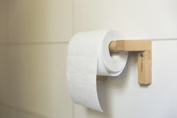 A white roll of soft toilet paper neatly hanging on a modern wooden holder on a white tile bathroom wall.