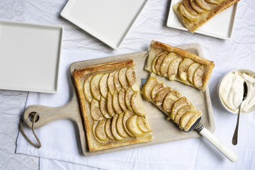 An apple tart cut into pieces on a wooden serving board, with plates and a bowl of cream.